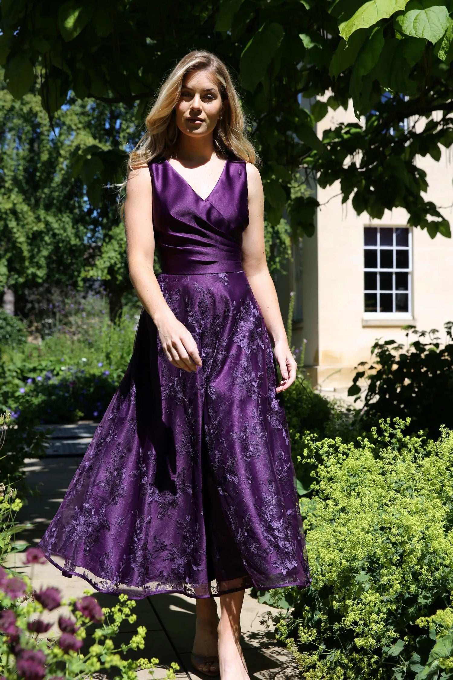 A woman wearing a sleeveless, midi-length blackberry purple dress with embroidery on the skirt.