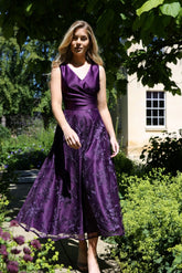 A woman wearing a sleeveless, midi-length blackberry purple dress with embroidery on the skirt.