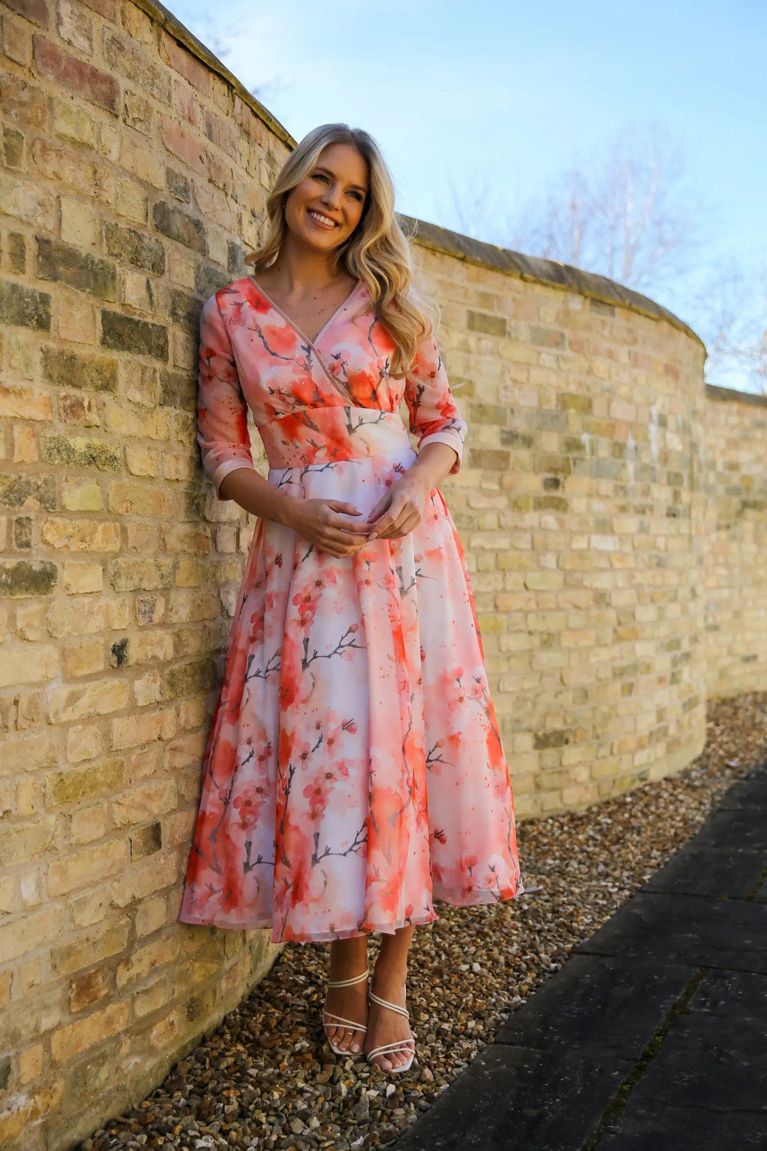 A woman standing against a brick wall wearing a pink floral chiffon occasion dress with sleeves and satin trim at the neck and wrists.