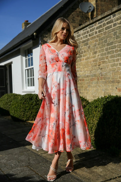 A woman standing against a brick wall wearing a pink floral chiffon occasion dress with sleeves and satin trim at the neck and wrists.