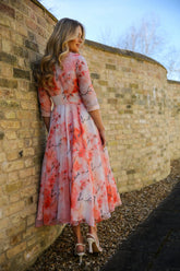 A woman standing against a brick wall wearing a pink floral chiffon occasion dress with sleeves and satin trim at the neck and wrists.