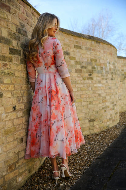 A woman standing against a brick wall wearing a pink floral chiffon occasion dress with sleeves and satin trim at the neck and wrists.