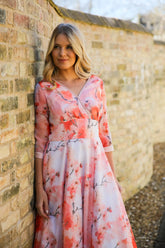 A woman standing against a brick wall wearing a pink floral chiffon occasion dress with sleeves and satin trim at the neck and wrists.
