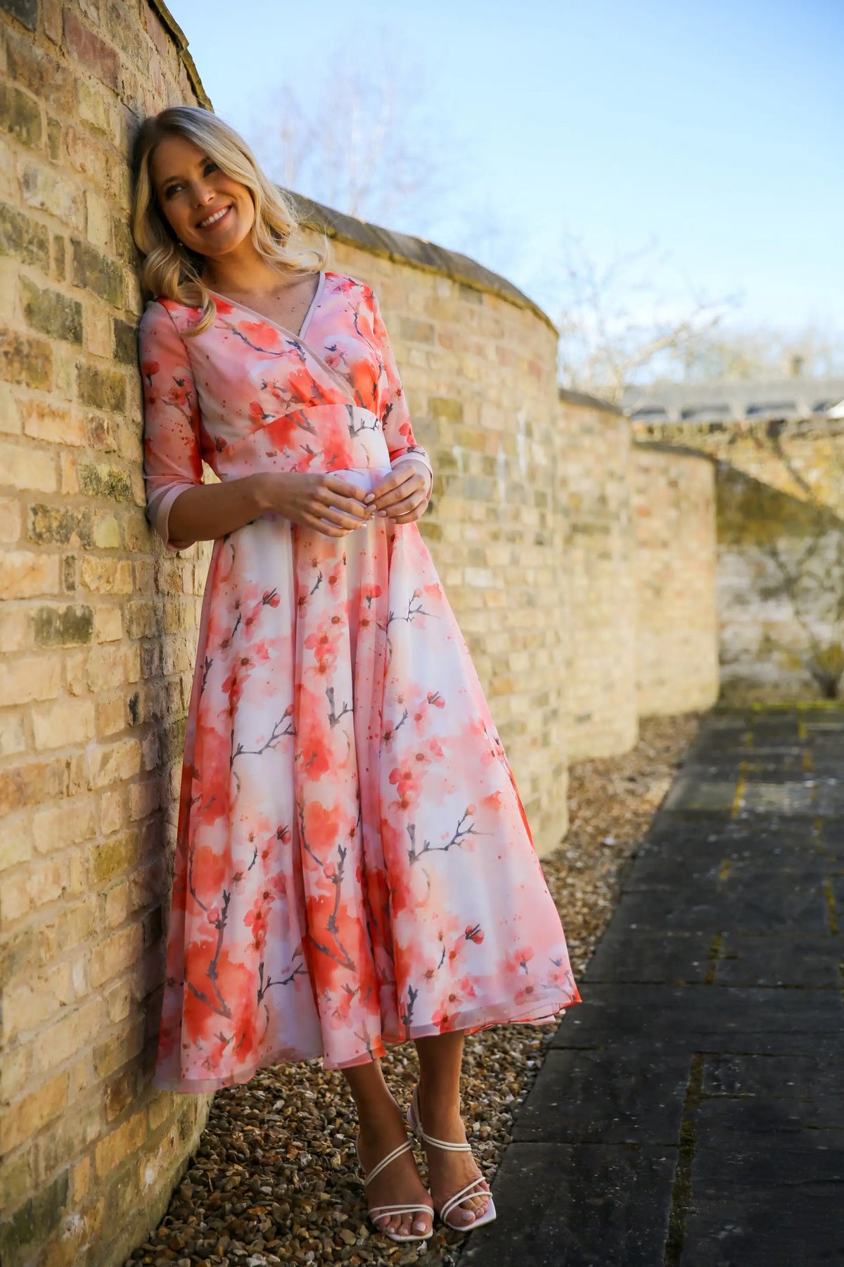 A woman standing against a brick wall wearing a pink floral chiffon occasion dress with sleeves and satin trim at the neck and wrists.