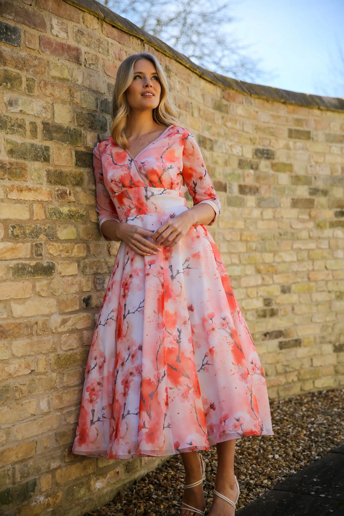 A woman standing against a brick wall wearing a pink floral chiffon occasion dress with sleeves and satin trim at the neck and wrists.