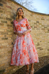 A woman standing against a brick wall wearing a pink floral chiffon occasion dress with sleeves and satin trim at the neck and wrists.