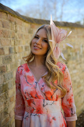 A woman standing against a brick wall wearing a pink floral chiffon occasion dress with sleeves and satin trim at the neck and wrists.