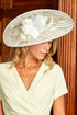 A woman wearing a pale green wrap dress and a large green fascinator hat with white floral accents, posed in front of a dark wood panelled door.