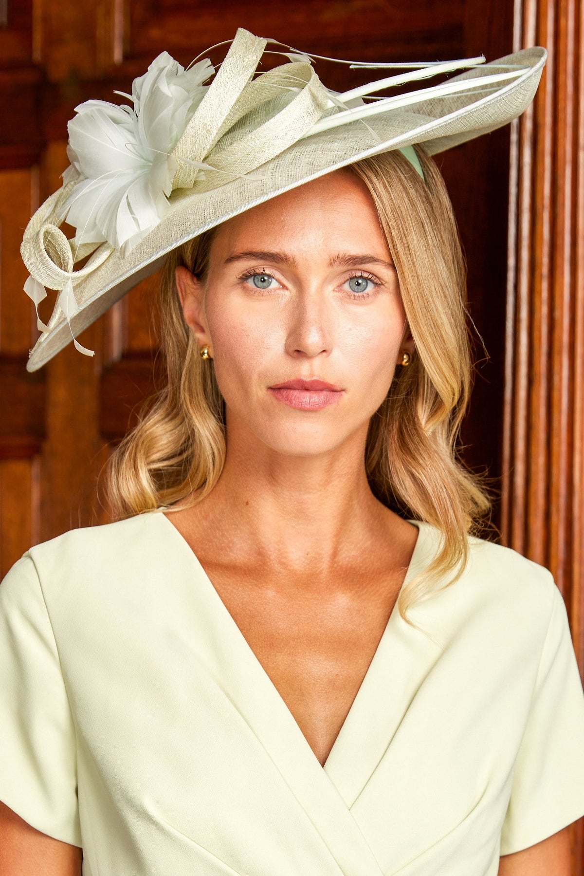 A woman wearing a pale green wrap dress and a large green fascinator hat with white floral accents, posed in front of a dark wood panelled door.