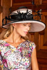 A woman wearing a floral print dress and a large black fascinator hat with feather accents, posed in front of a warm wood-paneled background.