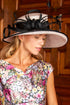 A woman wearing a floral print dress and a large black fascinator hat with feather accents, posed in front of a warm wood-paneled background.