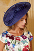 A woman wearing a large navy blue fascinator hat and a cream-colored dress with a colorful floral print, posed against a warm beige background.