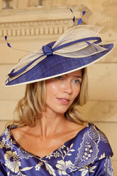 A woman wearing a wide-brimmed blue and beige fascinator hat with matching blue and beige floral dress, posed against a beige stone background.