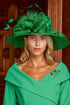 A woman wearing a bright green wide-brimmed fascinator hat with matching green dress, posed in front of a dark wood-panelled background.