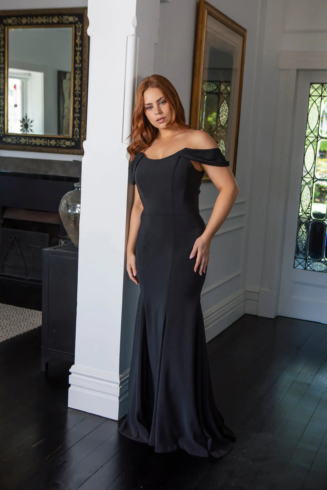 Woman in a black off-shoulder gown standing in a room with mirrors and a door.