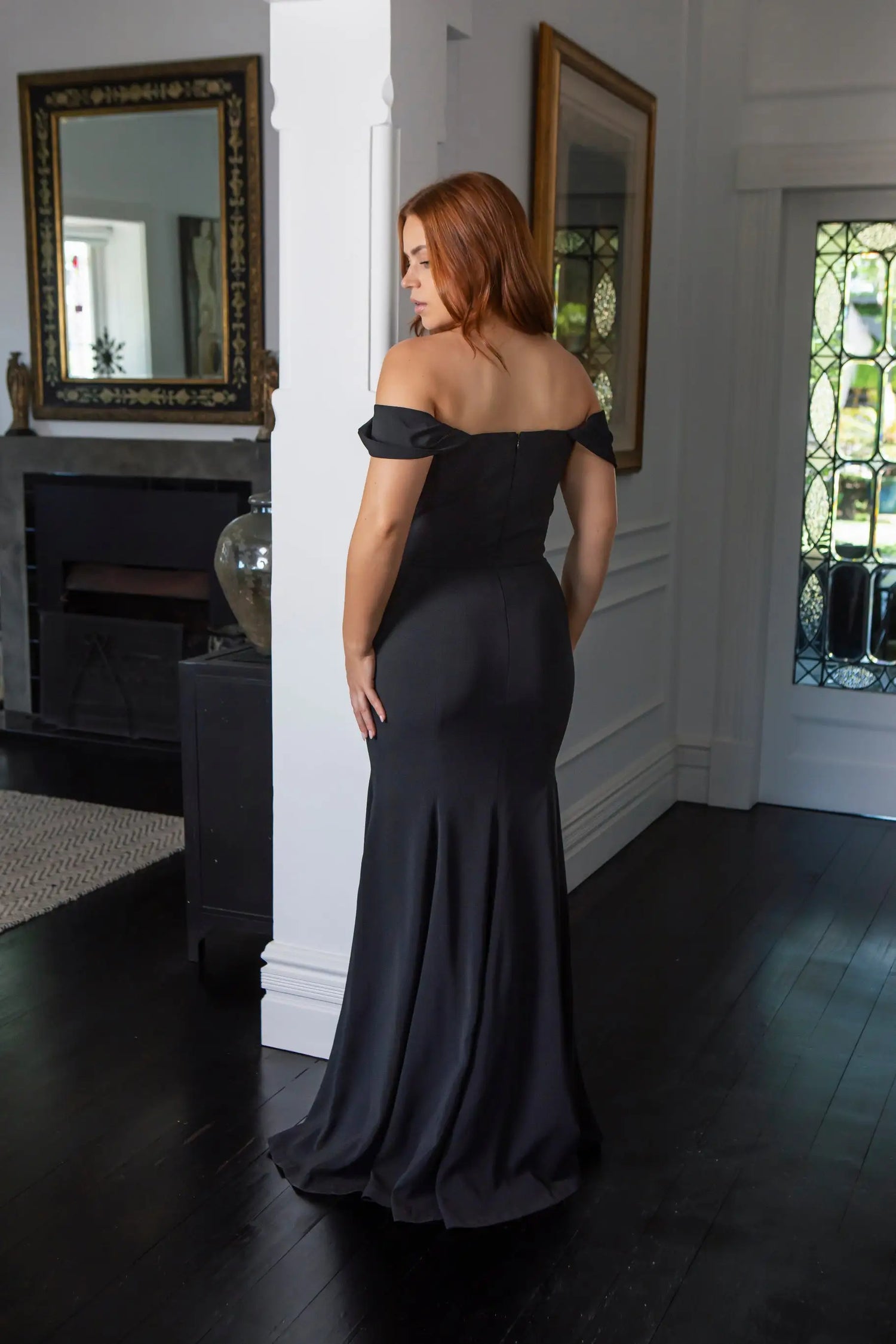 Woman in a black off-shoulder gown standing in a room with mirrors and a fireplace.