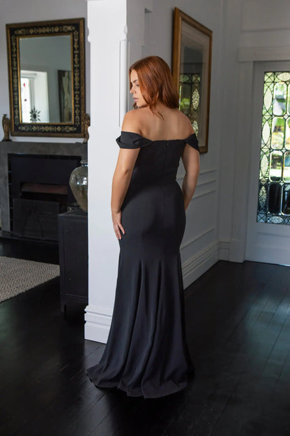 Woman in a black off-shoulder gown standing in a room with mirrors and a fireplace.