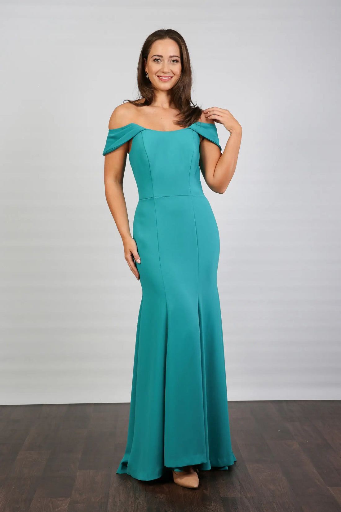 A woman in an off-the-shoulder turquoise maxi dress with a fit and flare silhouette, made from crepe fabric, posed against a neutral background.