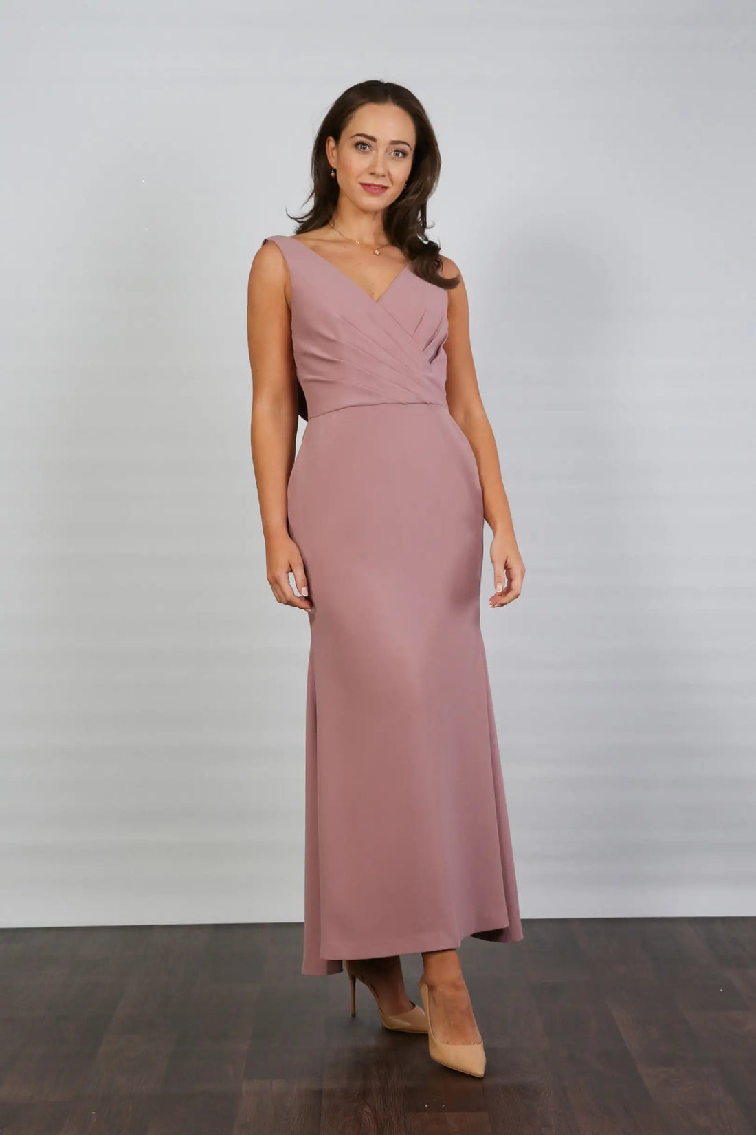 A model wearing a sleeveless, V-neck, dusty rose maxi dress with a deep cowl back and pleated waist, suitable for day to evening events.