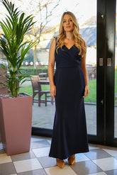 A woman wearing a navy maxi dress with a deep cowl back and pleated waist, with the dress ankle length. 
