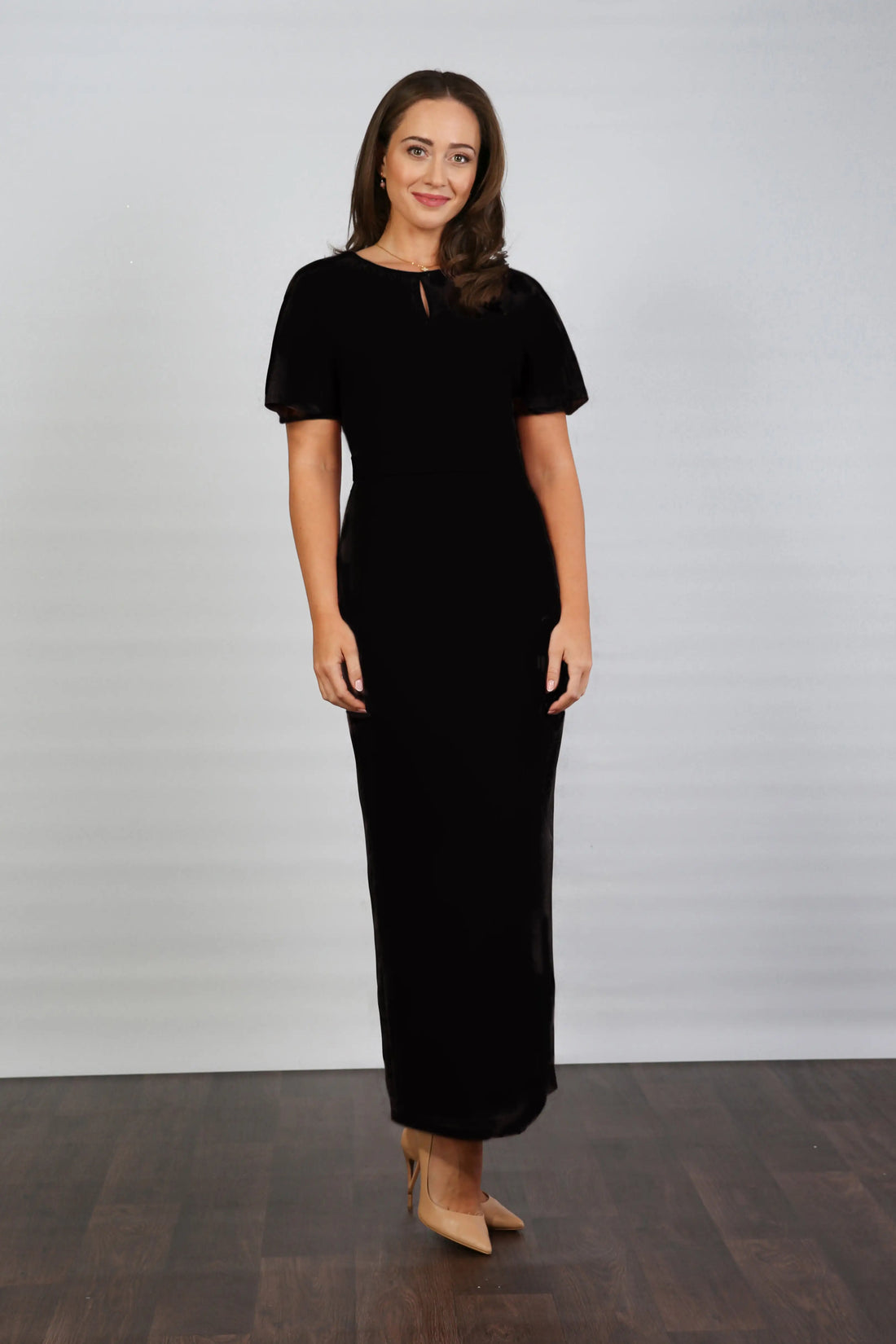 A woman models an ankle-length black dress with short sleeves and a V-neckline, standing against a plain background.