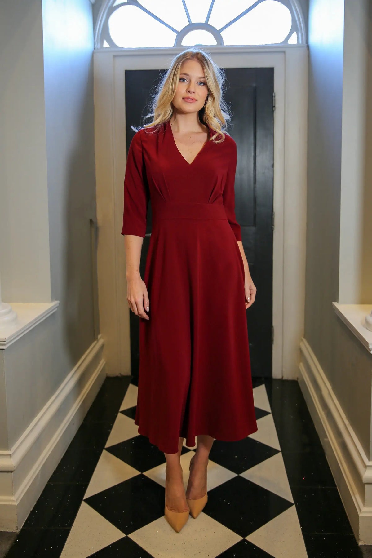 A model wearing a red, midi-length dress with a V-neck and 3/4 sleeves, standing in a room with black and white tiled floor.