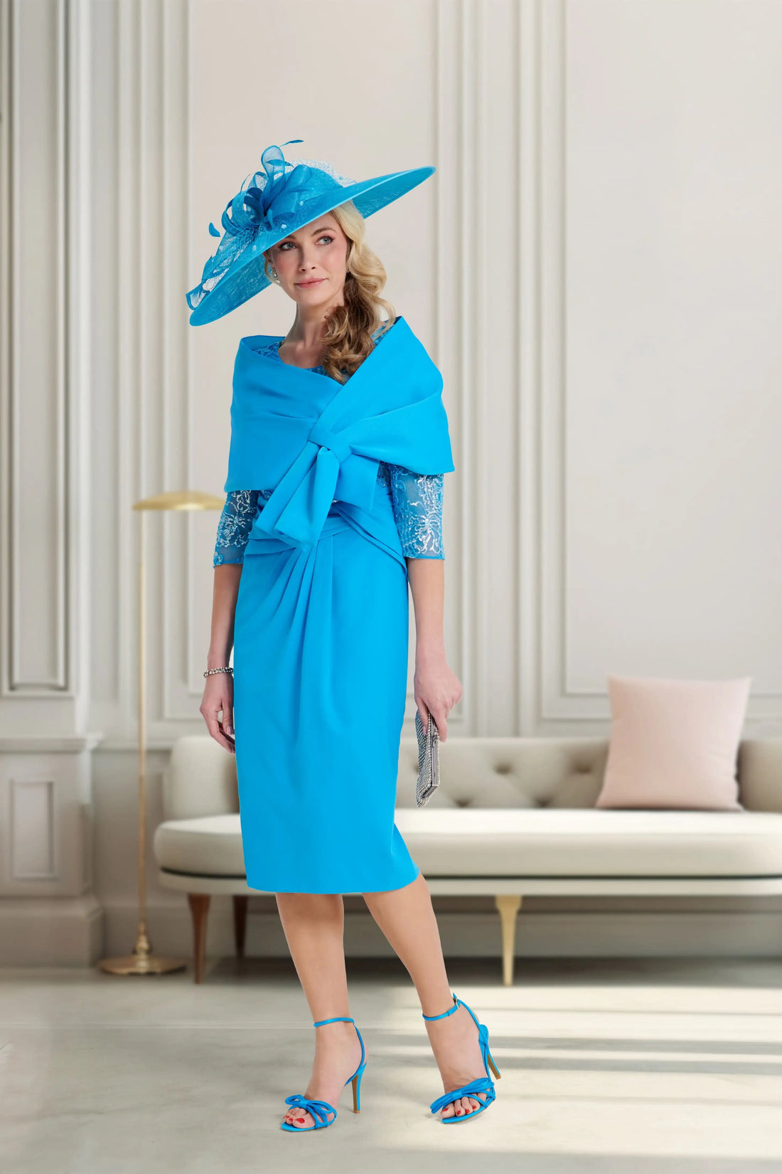 Woman in a blue dress and hat standing in a stylish room with a sofa and decorative pillows.