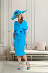 Woman in a blue dress and hat standing in a stylish room with a sofa and decorative pillows.