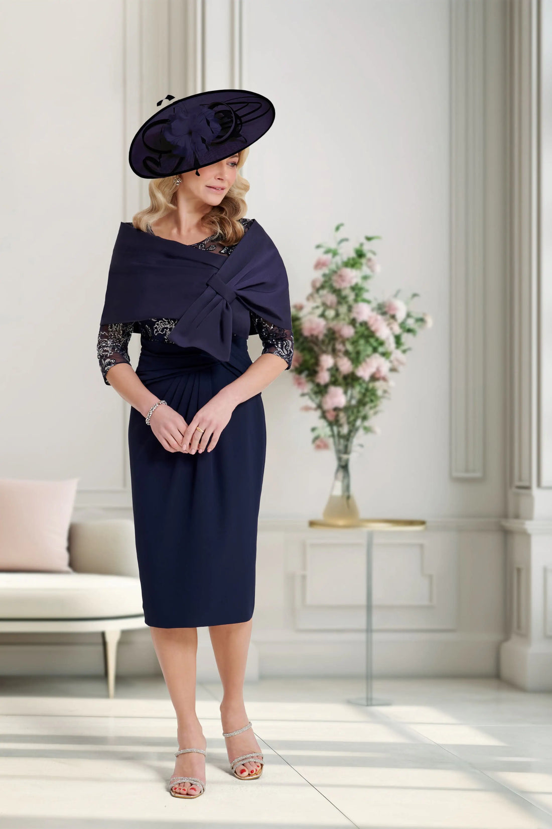 Woman in a navy dress with a matching hat standing in a well-lit room with a vase of flowers.