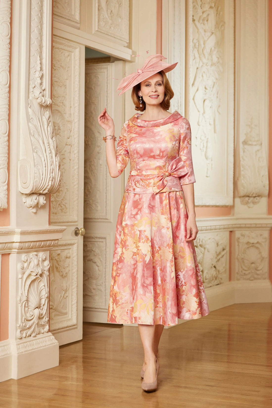 Woman in a floral dress and hat standing in an ornate room.