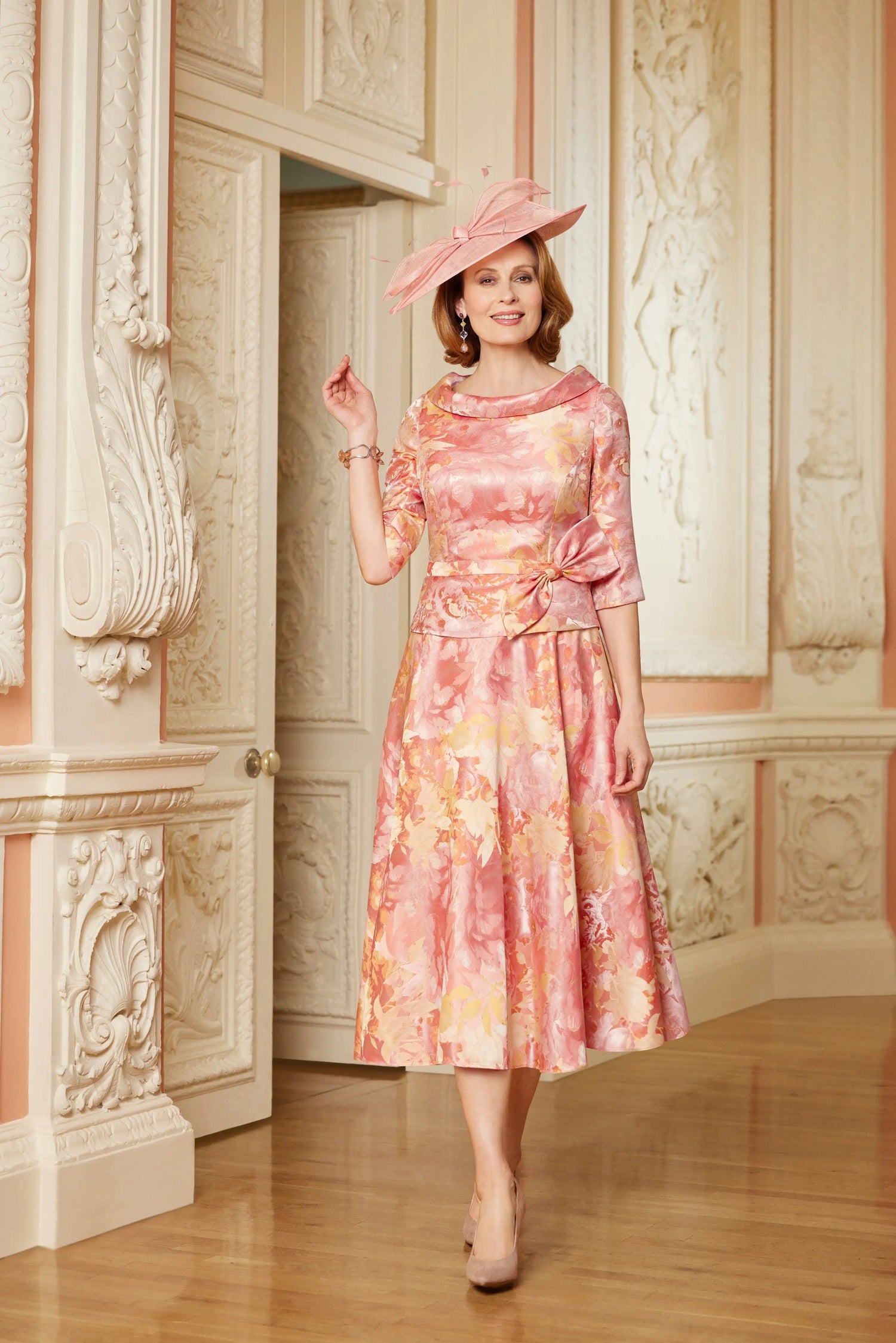 Woman in a floral dress and hat standing in an ornate room.