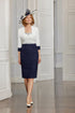 Woman wearing a white top with long sleeves, navy skirt, and matching shoes in a room with white walls.