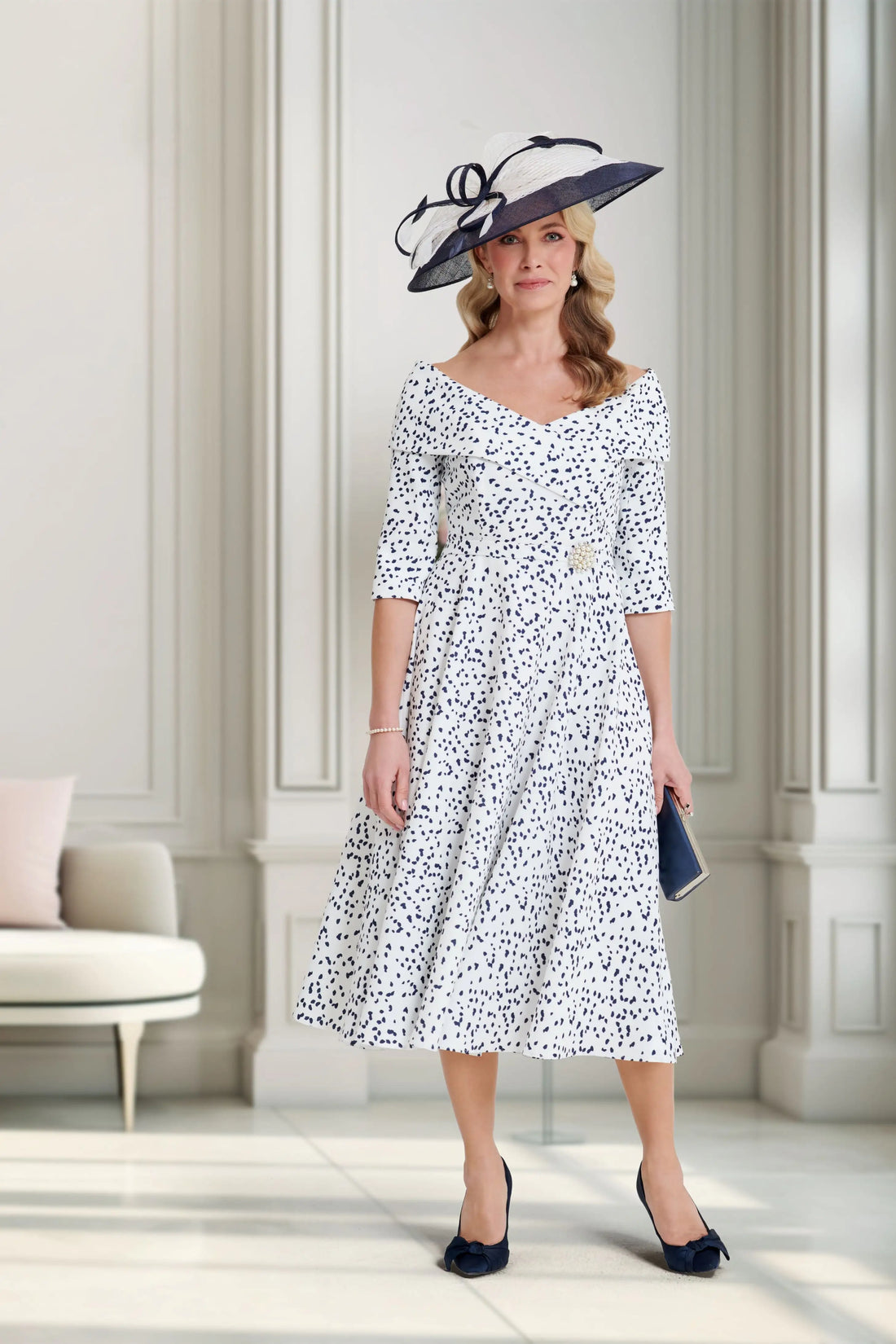 Woman in a white floral dress with a black hat standing in a bright room.