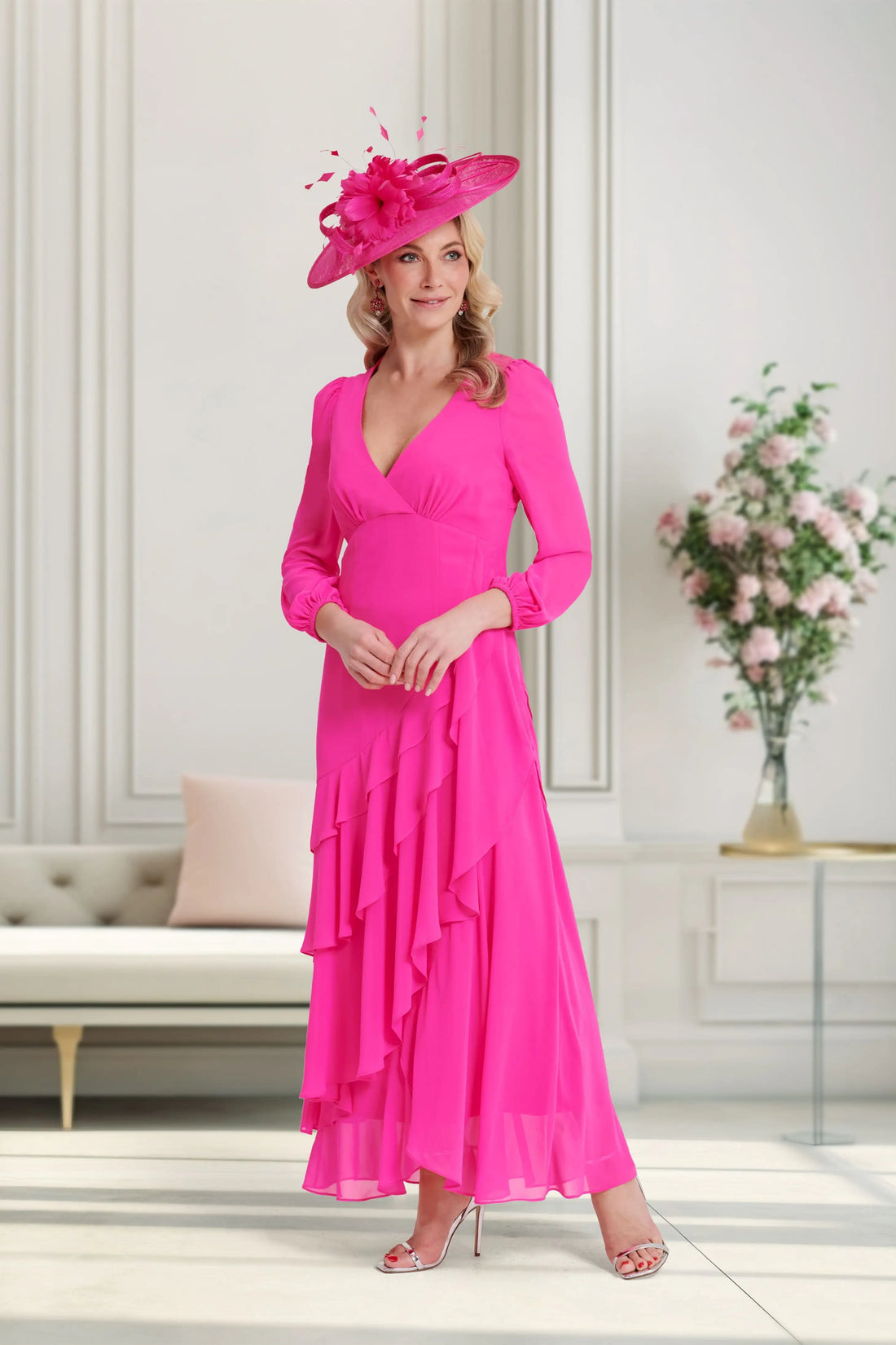 Woman in a bright pink dress and hat standing in a stylish room with a couch and floral arrangement.