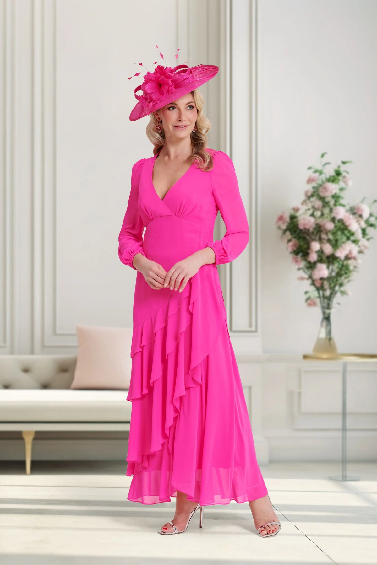Woman in a bright pink dress and hat standing in a stylish room with a couch and floral arrangement.