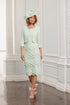 A woman wearing a light green scallop lace occasion dress with sleeves and a matching bolero jacket, complete with a hat, shoes, and a bow detail.