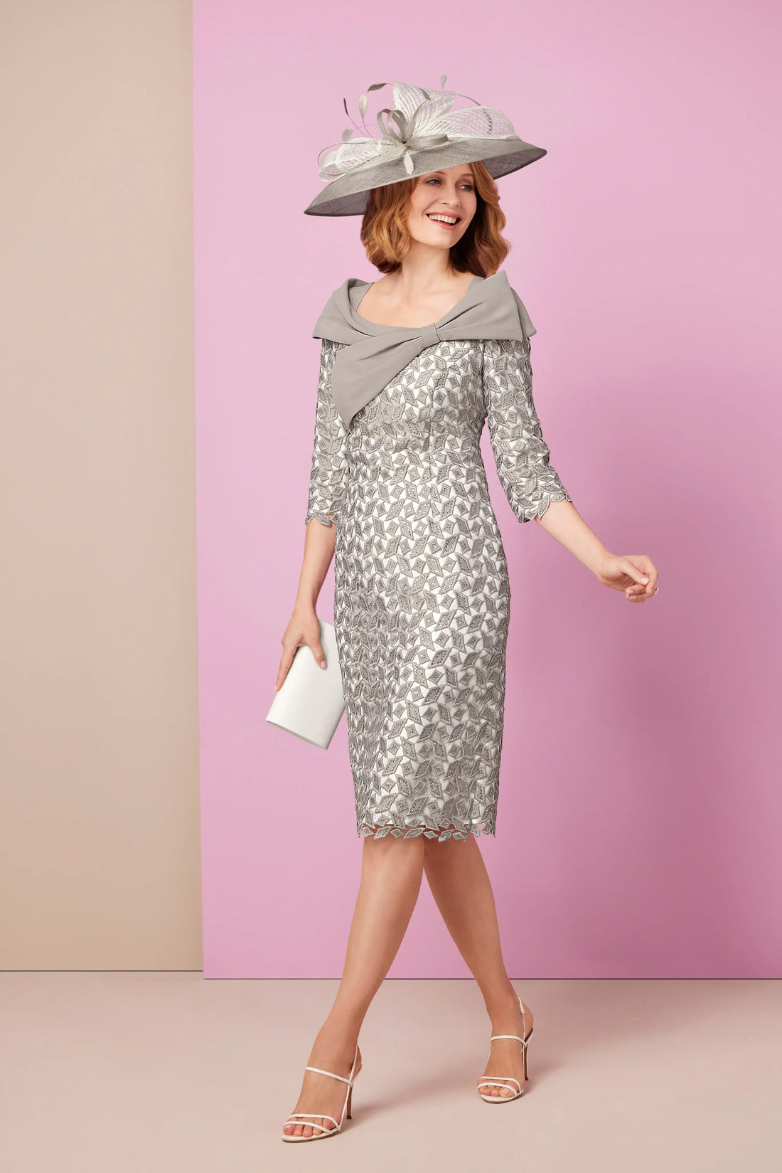 A woman wearing a silver long sleeved knee length dress with a bow detail, paired with a hat and heels.