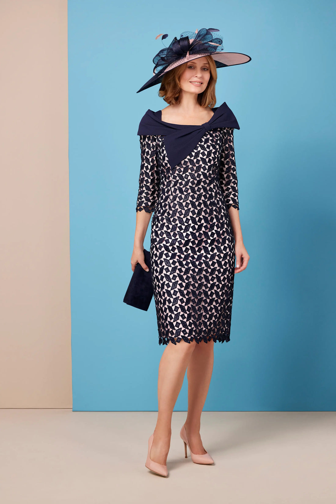 A model wearing a navy shift silhouette dress with a geometric pattern, featuring long sleeves and a bow detail on the neckline. The dress is fully lined and finishes below the knee.