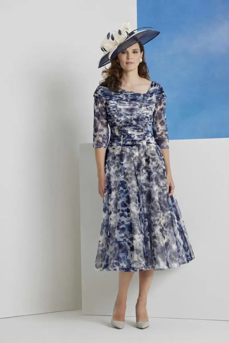 Woman wearing a floral dress and large hat against a white wall with blue sky.