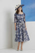 Woman wearing a floral dress and large hat against a white wall with blue sky.