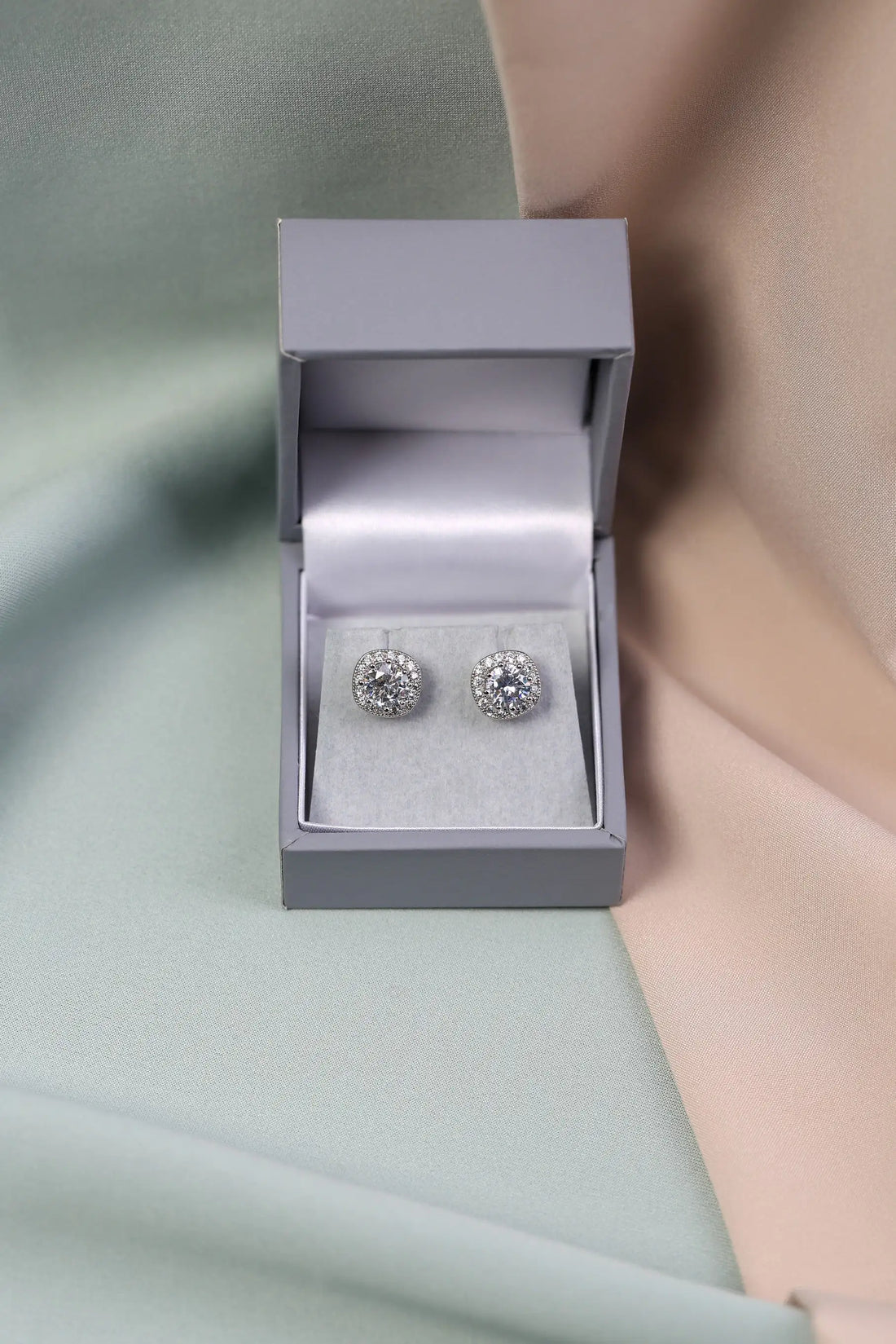 A pair of crystal halo stud earrings displayed in an open grey box, with the earrings themselves having a large central clear crystal surrounded by smaller clear crystals.