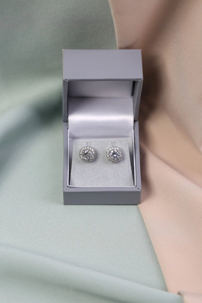 A pair of crystal halo stud earrings displayed in an open grey box, with the earrings themselves having a large central clear crystal surrounded by smaller clear crystals.