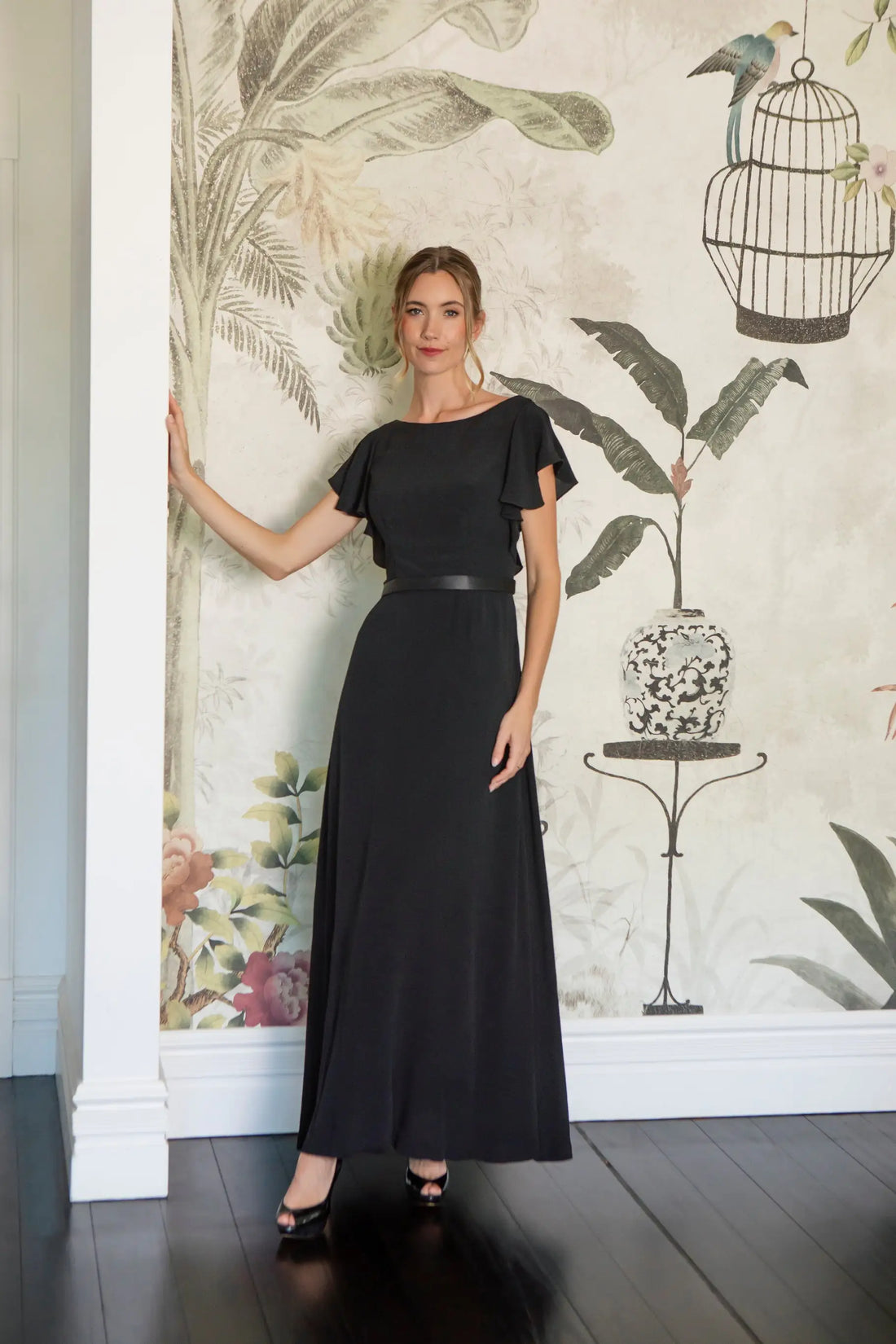 Woman in a black dress standing against a decorative wall with floral and birdcage designs.