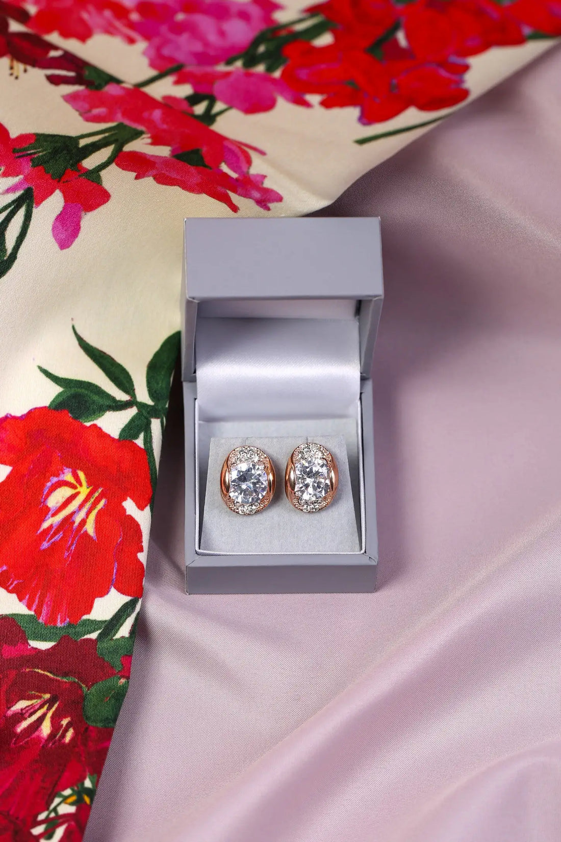 A pair of crystal earrings in a gold setting, presented in an open luxury box on a floral patterned fabric.