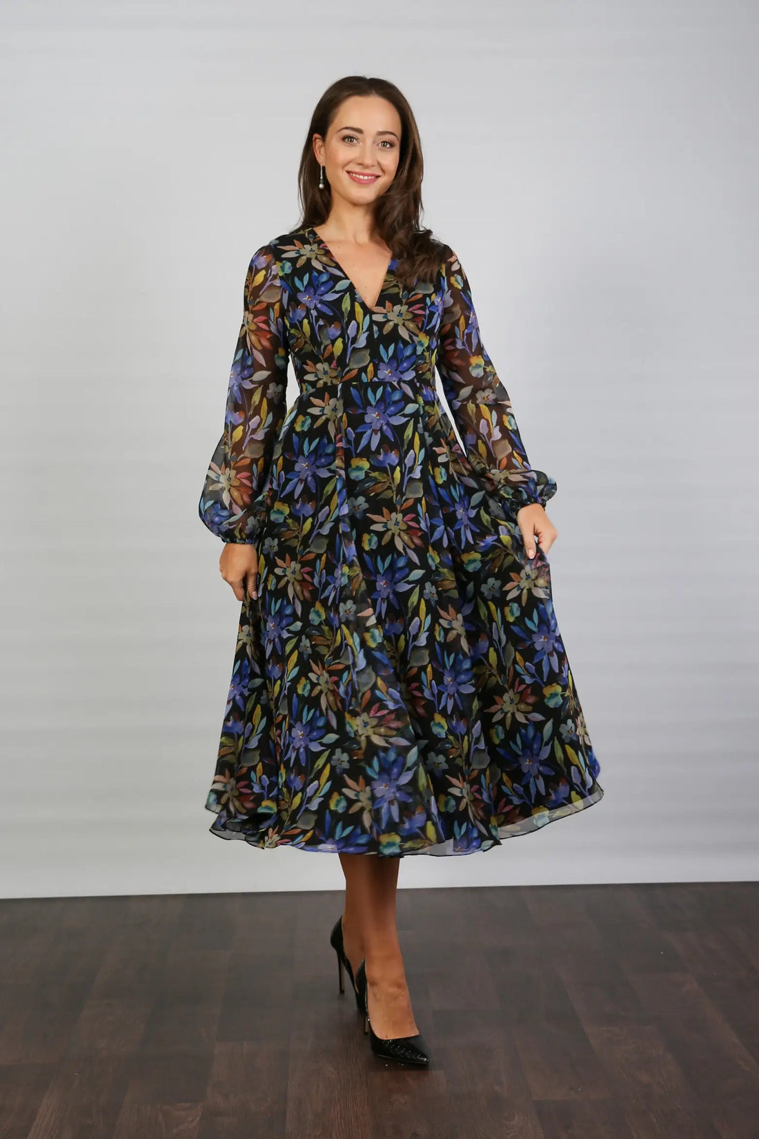 A woman standing indoors wearing a floral printed chiffon midi dress with long sleeves and a round neckline.