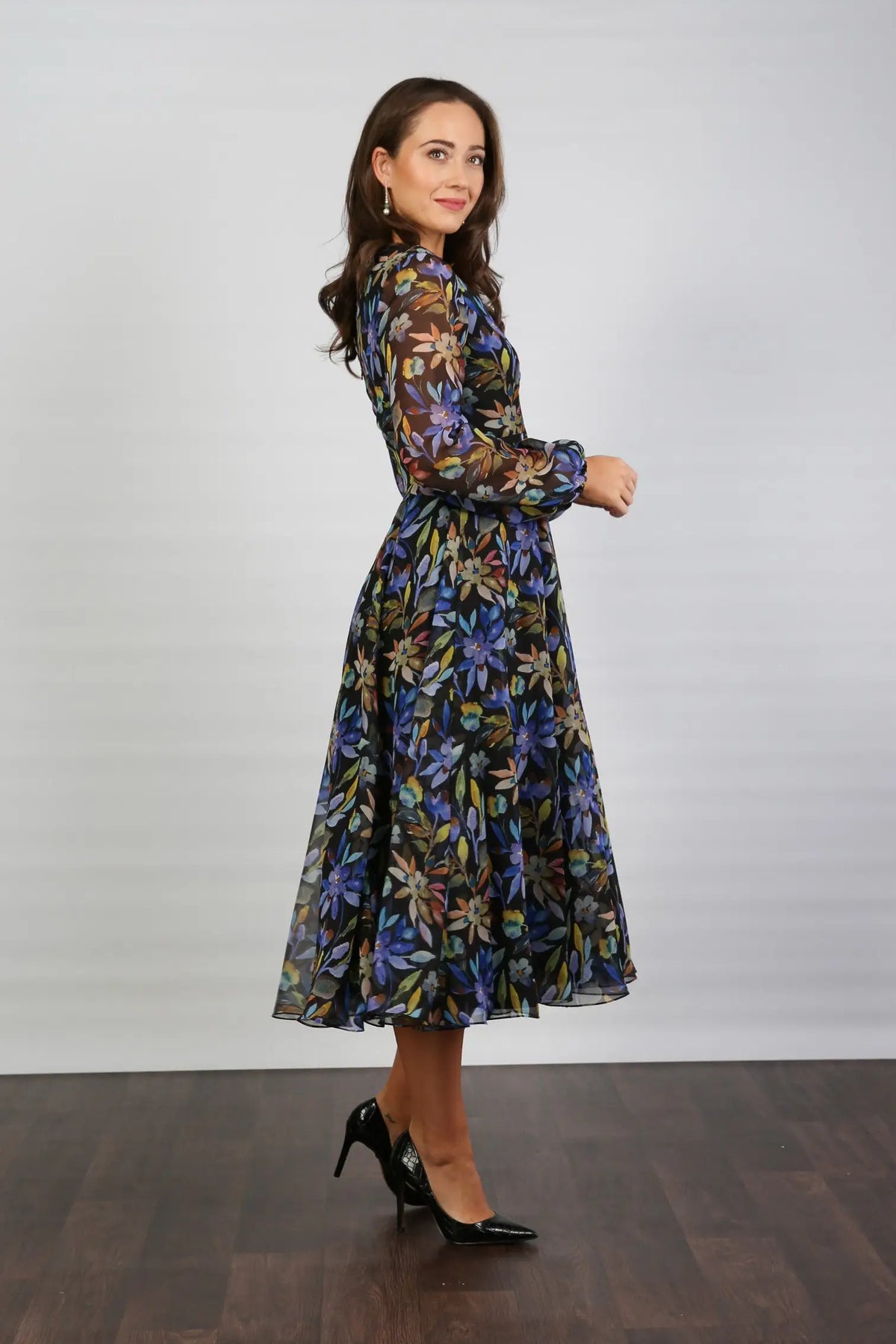 A woman standing indoors wearing a floral printed chiffon midi dress with long sleeves and a round neckline.