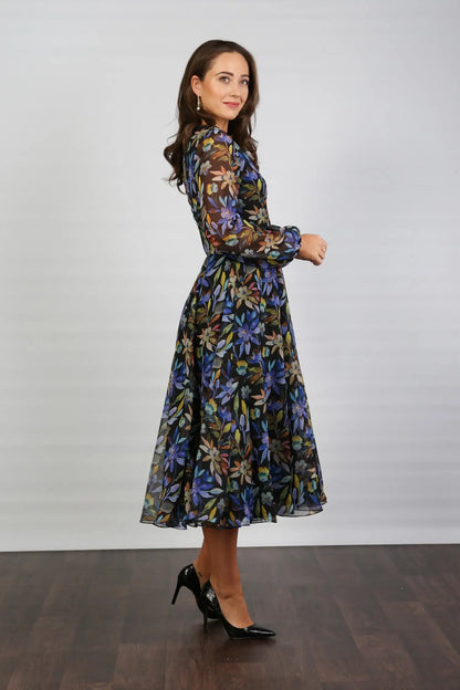A woman standing indoors wearing a floral printed chiffon midi dress with long sleeves and a round neckline.