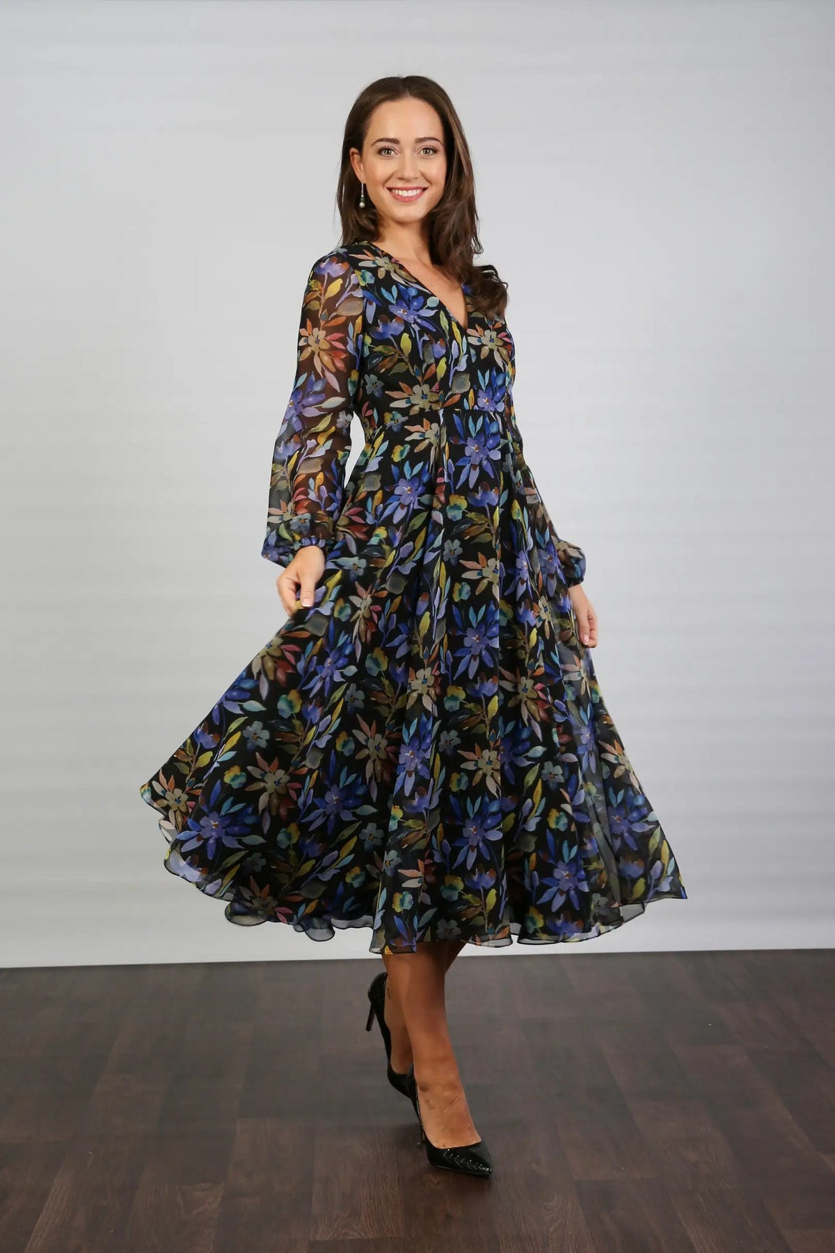 A woman standing indoors wearing a floral printed chiffon midi dress with long sleeves and a round neckline.