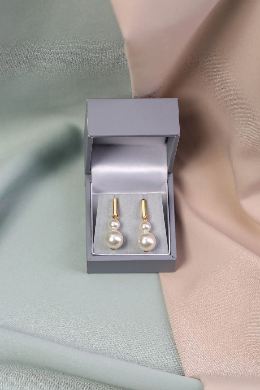 A pair of gold bar pearl drop earrings displayed in an open grey box on a silky dual-tone background.