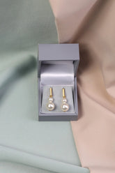 A pair of gold bar pearl drop earrings displayed in an open grey box on a silky dual-tone background.